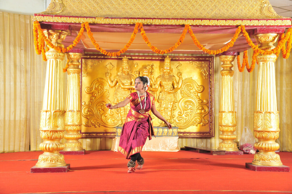 Photo of Priyanka Saminathan in traditional Indian dance costume. Performing on a red stage with a golden roof and gold pillars.