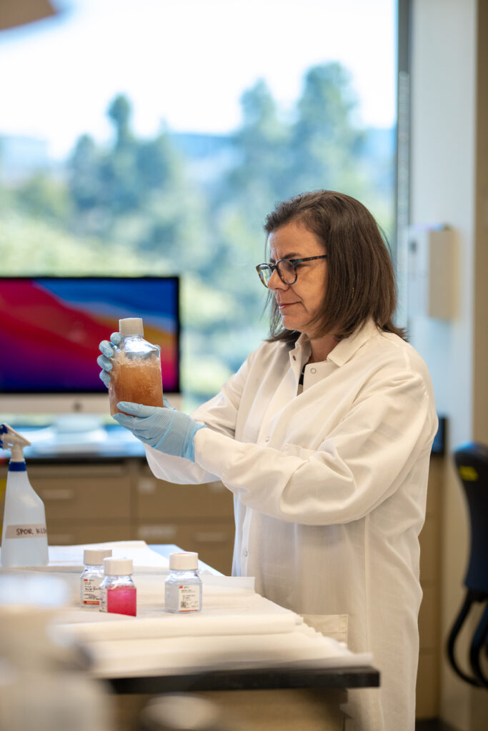 Bianca Mothé, Ph.D., in a lab setting. Wear a white coat and hold a sample in a flask