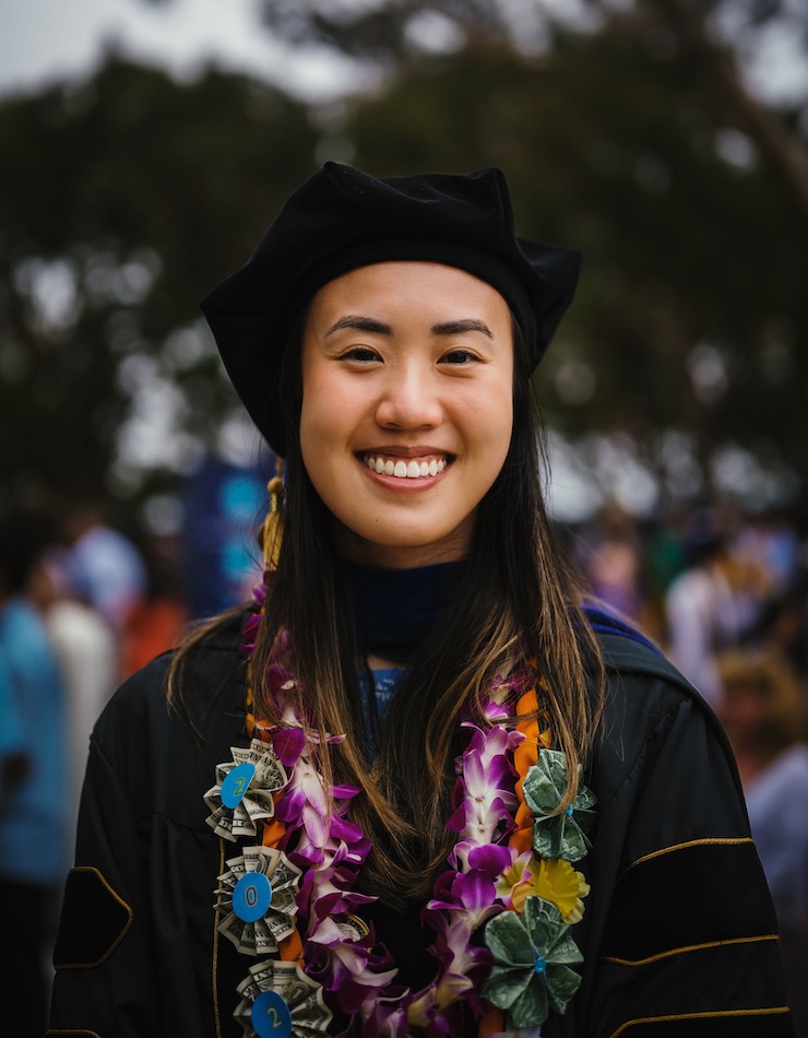 Ivy Phung faces the camera and smiles. She is wearing a black graduation robe and cap. She is wearing a necklace of flowers and a necklace of dollar bills.