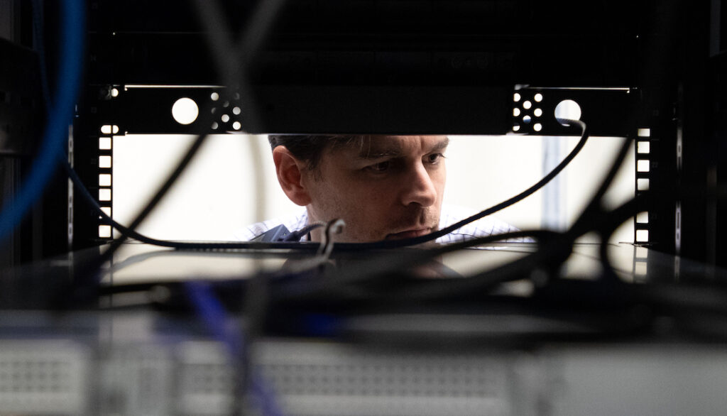 Michael Scarpelli is shown looking through a computer server area. His face is framed by machinery and wires.