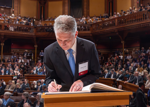 Shane Crotty writes his name in a large book. He is wearing a suit. There is a crowd of people behind him.