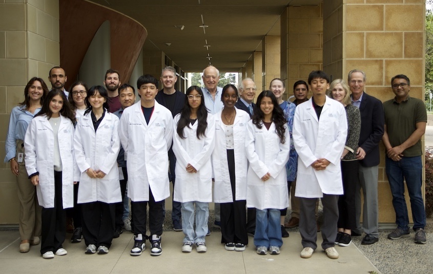 A group of students in white lab coats stand in front of a row of LJI employees and Institute supporters