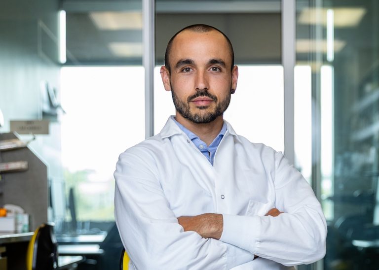Portrait photo of Dr. Reina-Campos. He is sitting in his laboratory. He is wearing a white lab coat