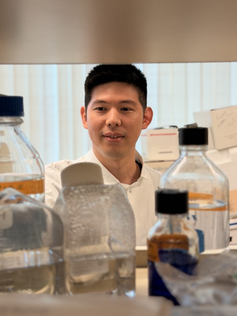 Photo of Dr. Lee in the lab. He is wearing a white lab coat and is surrounded by bottles and lab supplies